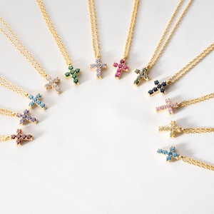 May include: A collection of gold-toned cross necklaces, each adorned with colorful gemstones. The crosses feature various jewel tones, including blue, green, pink, and purple. The necklaces are arranged in a semi-circle on a white background.