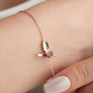 May include: A rose gold chain bracelet featuring a hummingbird charm. The hummingbird is accented with a pear-shaped red gemstone in the center and small green stones embedded along the wings and head, creating a detailed avian design.