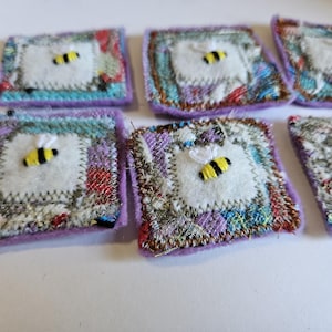 May include: A collection of handmade fabric squares with a bee applique. Each square features a white felt center with a yellow and black bee, surrounded by colorful patchwork and a lavender border. The squares are arranged on a white surface.