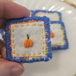 May include: A square patch featuring a white felt center with an embroidered orange pumpkin and a green stem. The patch is bordered by a thin orange layer and a thick blue blanket-stitched edge, with yellow running stitches around the pumpkin.
