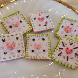 May include: Close-up of six square felt patches with embroidered hearts and decorative stitching. The patches are white with pink and purple accents, and green stitching around the edges. The patches are arranged on a white plate.