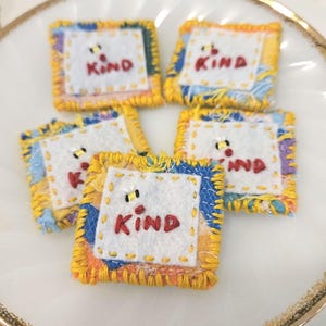 May include: Five square fabric patches featuring white centers with the embroidered red text "KIND" and a small bee icon. Each patch has a yellow stitched border and is layered over a multi-colored fabric base with frayed edges and yellow thread.