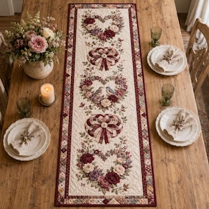 May include: A rectangular quilted table runner featuring a cream-coloured background with a repeating pattern of floral hearts, woven ribbon hearts, and two small birds. The runner has a dark red border and is decorated with intricate stitching.