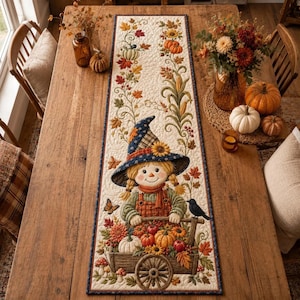Puede incluir: Un camino de mesa acolchado rectangular con una ilustración central de un espantapájaros con sombrero azul, de pie detrás de un carro de madera lleno de calabazas, girasoles y hojas de otoño, acentuado por un pájaro negro.