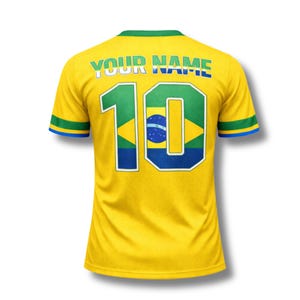 Może przedstawiać: Ż&oacute;łta koszulka piłkarska Brazylii z zielonymi i niebieskimi akcentami. Numer 10, zawierający flagę Brazylii, jest wyświetlany na plecach. Tekst "YOUR NAME" jest łukowato nad numerem.