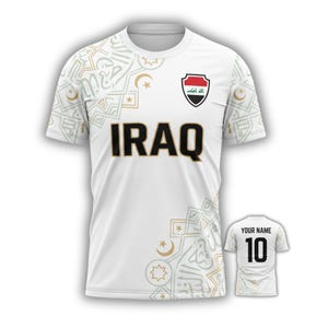 May include: White soccer jersey with the word "IRAQ" in black and gold lettering. The jersey features a crest with the Iraqi flag and decorative patterns. A smaller jersey shows "YOUR NAME" and the number "10".
