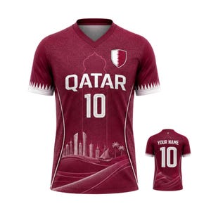 Puede incluir: Camiseta de f&uacute;tbol granate con detalles blancos, que muestra la palabra "QATAR" y el n&uacute;mero "10" en blanco. La camiseta tiene cuello en V y un gr&aacute;fico de horizonte. Una versi&oacute;n m&aacute;s peque&ntilde;a de la camiseta se muestra a la derecha.