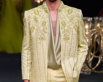 Könnte beinhalten: Ein hellgelber Anzug mit aufwendiger floraler Stickerei und Perlenverzierungen. Die Jacke hat einen Stehkragen und wird mit einer passenden Hose kombiniert. Dazu ein hellbeige Hemd. Der Stil ist formell und elegant.