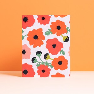 May include: A rectangular card featuring a repeating pattern of stylized orange and red poppies with black centers and green leaves on a white background. The card is standing upright against an orange backdrop.