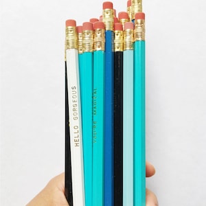 May include: A collection of hexagonal pencils in shades of white, black, and various tones of blue. Each pencil features a gold-colored metal ferrule and a pink eraser. Two pencils display gold foil text reading HELLO GORGEOUS and YOU'RE MAGICAL.