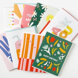 May include: A collection of folded greeting cards featuring various graphic designs, including a green card with floral patterns and the text HAPPY BIRTHDAY, a card with the text THANK YOU, and others with colorful stripes, fruit motifs, and text.