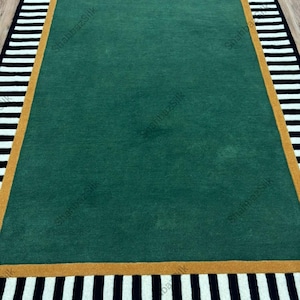 May include: A rectangular area rug featuring a solid deep green center field, bordered by a thick mustard yellow frame, and finished with an outer edge of alternating black and white vertical stripes. The text "ShabbazSilk" is visible.