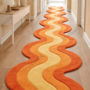 May include: A long, wavy rug in shades of orange and yellow, creating a retro design. The rug is placed in a hallway with light-coloured walls and wooden flooring. The rug's dimensions are not visible.