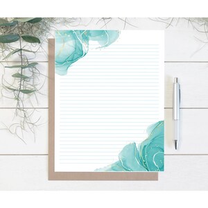 May include: A lined stationery set featuring a white paper with teal watercolor accents and gold flecks. A silver pen rests on the right side. The stationery is on a brown backing, with greenery in the background.