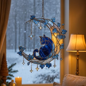Könnte beinhalten: Glasfenster-Hängeornament mit einer blauen Katze auf einem Mond. Die Katze hat ein Sternenmuster und sitzt auf einem gelb-blauen Mond. Blaue Blätter und hängende Sterne schmücken den Mond. Das Fenster zeigt eine verschneite Landschaft.