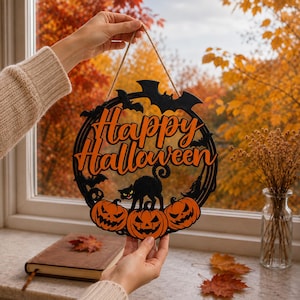 May include: A circular black and orange wooden Halloween sign featuring the text Happy Halloween in orange script. The design includes a black cat, three orange jack-o'-lanterns, and black bats. The sign hangs from a thin brown twine string.