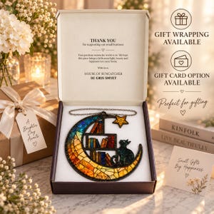 May include: A stained glass suncatcher shaped like a crescent moon with a black cat sitting on a bookshelf, presented in an open gift box. The moon features orange, yellow, and blue glass segments, accompanied by a small star and a hanging chain.