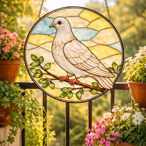 May include: A round stained glass window featuring a white dove perched on a branch with green leaves. The background is a mosaic of yellow, blue, and white glass. The window is hanging from a gold chain.