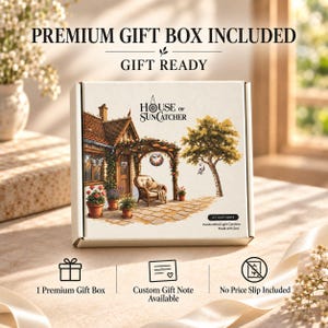May include: A white square cardboard gift box featuring the text HOUSE OF SUNCATCHER and an illustration of a cottage patio with a wicker chair, potted plants, and a tree. The box includes text stating PREMIUM GIFT BOX INCLUDED and GIFT READY.