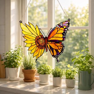 May include: A stained glass butterfly suncatcher, half sunflower and half monarch butterfly, hangs in front of a window. The butterfly features yellow, orange, purple, and black colors. The suncatcher is suspended by a silver chain.