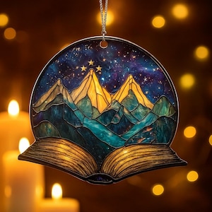 Peut inclure: Ornement rond en vitrail représentant une chaîne de montagnes sous un ciel étoilé. Les montagnes sont jaunes et bleues, avec un ciel bleu foncé et violet. L'ornement a la forme d'un livre ouvert et est suspendu à une chaîne argentée.