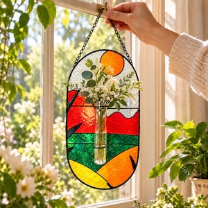 May include: An oval stained glass wall hanging featuring a landscape of red, green, and yellow sections with a circular orange sun. A clear glass test tube vase is attached to the center, holding a bouquet of white flowers and green leaves.