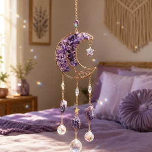 Può includere: Un ornamento sospeso con una cornice a mezzaluna color oro riempita di piccole scaglie di ametista viola e un design ad albero in filo metallico. Tre catenelle dorate pendono dalla luna, decorate con pietre di ametista e sfere di cristallo.