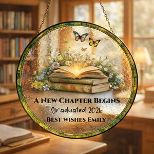 May include: Round stained glass panel with a green border, featuring a stack of books with an open, glowing book on top. Butterflies and flowers surround the books. Text reads "A NEW CHAPTER BEGINS Graduated 2026 BEST WISHES EMILY."
