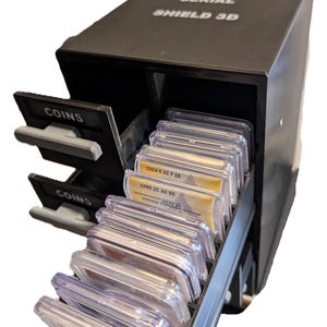 May include: Black coin storage box with the text "SERIAL SHIELD 3D" and "COINS". The box has multiple drawers filled with graded coin holders. The holders contain coins with labels visible, including "1924-5 1C F 15" and "1990 1C AU 55".
