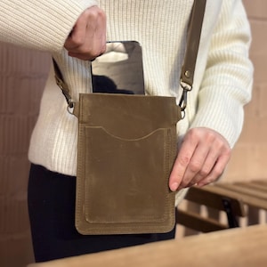 May include: A brown leather crossbody phone bag with a front pocket and adjustable strap. The bag is being held by a person wearing a cream-coloured sweater. The bag is designed to hold a smartphone.