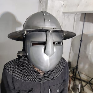 Puede incluir: Un casco medieval plateado con visera y ala ancha. El casco tiene remaches y se usa con una cofia de cota de malla. La persona lleva una camisa negra.