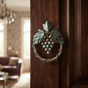 Puede incluir: Un llamador de puerta de metal con un diseño de anillo circular envuelto en metal con forma de enredadera, rematado con hojas en tonos verdes y un racimo de acentos esféricos iridiscentes similares a uvas, montado en una puerta de madera oscura.