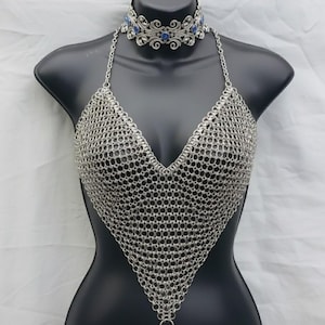 Puede incluir: Top halter de cota de malla plateada con cuello en V profundo y collar gargantilla a juego. El collar presenta detalles plateados ornamentados y piedras preciosas azules. El top se exhibe en un maniquí negro.