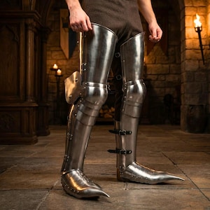 May include: A pair of polished steel leg armor pieces covering the thighs, knees, and lower legs. The armor features articulated metal plates, pointed sabatons at the feet, and black leather straps with metal buckles securing the lower leg sections.