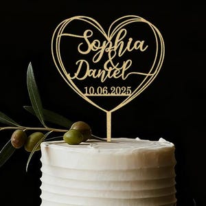 Peut inclure: Un gâteau de mariage blanc à deux étages avec un cake topper doré en forme de cœur. Le cake topper porte les noms "Sophia & Daniel" et la date "10.06.2025". Des branches d'olivier avec des olives vertes décorent le gâteau.