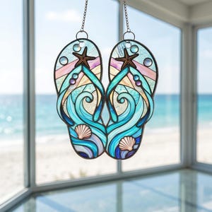 May include: Stained glass window decoration in the shape of flip-flops. The design features ocean waves, seashells, and starfish in shades of blue, teal, and purple. The flip-flops are suspended by chains, perfect for a beach-themed decor.