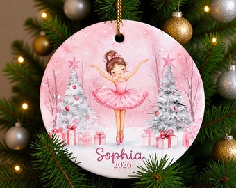 May include: A round ceramic Christmas ornament featuring a pink watercolour illustration of a ballerina in a tutu dancing between two white Christmas trees with pink gifts, personalised with the name Sophia and the year 2026 in pink text.