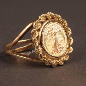 May include: A gold-toned ring featuring a coin with a detailed relief of a woman. The coin is encircled by a rope-like border, and the ring has a split shank design. The ring is set against a dark background.