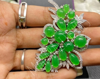 Pode incluir: Broche em forma de flor com pedras preciosas verdes vibrantes e detalhes claros e brilhantes. A broche tem uma base de metal prateado. As pedras preciosas são de vários tamanhos e formas, criando um design natural. Uma peça de joalharia marcante.