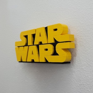 May include: A three-dimensional wall-mounted sign featuring the yellow text STAR WARS in a bold, blocky font. The letters have a layered, ridged texture and are mounted on a thin black backing plate, creating a distinct depth against the wall.