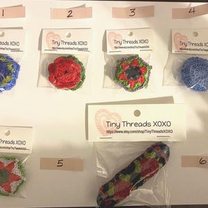 Może przedstawiać: Kolekcja ręcznie robionych szydełkowych przypinek w kształcie kwiatów w różnych kolorach, w tym niebieskim, czerwonym, zielonym i we wzór kwiatowy. Każda przypinka jest zapakowana z etykietą Tiny Threads XOXO.