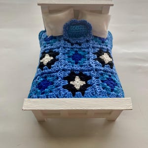 May include: A miniature white bed frame with a blue crocheted blanket. The blanket features a pattern of dark blue, light blue, black, and white squares. Two white pillows and a blue crocheted pillow are on the bed.