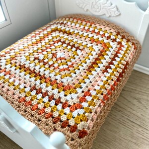 May include: A miniature white bed with a handmade crochet blanket. The blanket features a square pattern in shades of brown, orange, yellow, peach, and white. The bed frame has a decorative headboard.