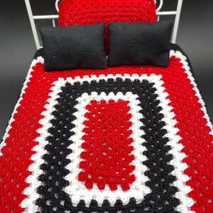 May include: A miniature white metal bed with a red, black, and white crocheted blanket. Two black pillows and a red crocheted pillow are on the bed. The bed is a dollhouse or miniature item.