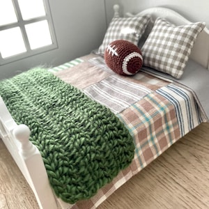 May include: A miniature bed with a patchwork quilt in neutral tones, a green knitted throw, and two checkered pillows. A brown and white crocheted football rests on the bed, suggesting a sports or nursery theme.