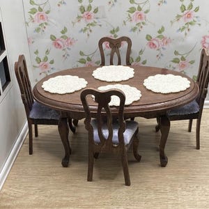 Puede incluir: Juego de comedor de madera en miniatura con una mesa redonda y cuatro sillas. La mesa es de color marrón oscuro con cuatro manteles individuales de encaje blanco. Las sillas tienen asientos tapizados grises. El fondo presenta papel pintado floral.