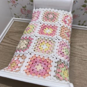 May include: A miniature white bed with a crocheted blanket. The blanket features a patchwork design with squares in shades of pink, yellow, and beige, bordered by white crochet. The bed frame is simple and painted white.