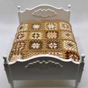 May include: A miniature white bed with a crocheted blanket. The blanket is made of squares in shades of brown, beige, and cream. The bed frame has decorative carvings on the headboard and footboard.