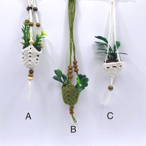 Op de afbeelding: Drie hangende macramé plantenhangers met kunstgroen. De houders zijn in wit en groen, met houten kralen en een wit koord. De planten staan in kleine, decoratieve potten. De houders zijn gelabeld met A, B en C.