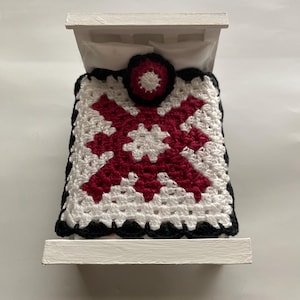 May include: A miniature white bed with a crocheted blanket and pillow. The blanket features a red and white geometric design with a black border. The pillow has a red and black floral design.